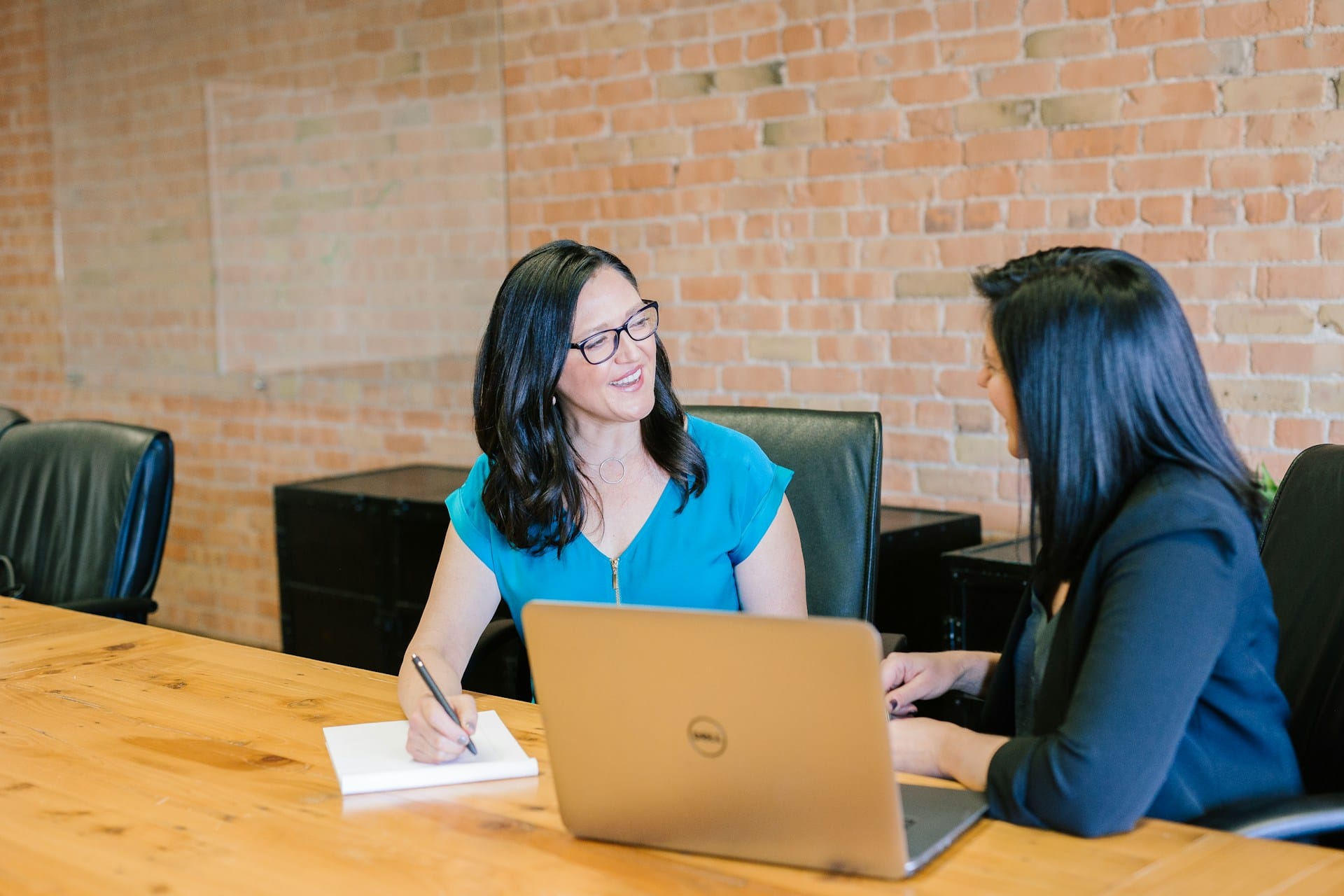 Two businesswomen having a friendly discussion at a wooden desk, one taking notes beside a laptop