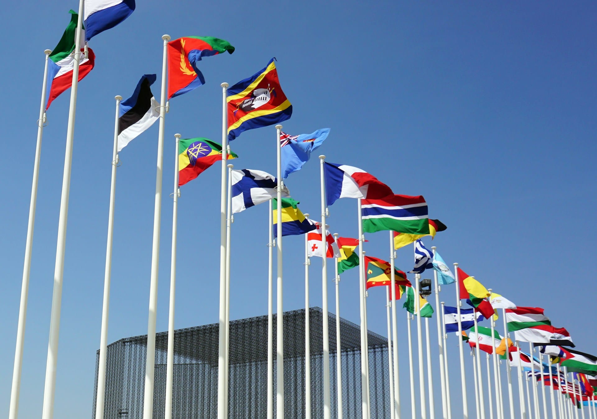 Row of international flags flying against a clear blue sky outside a modern building
