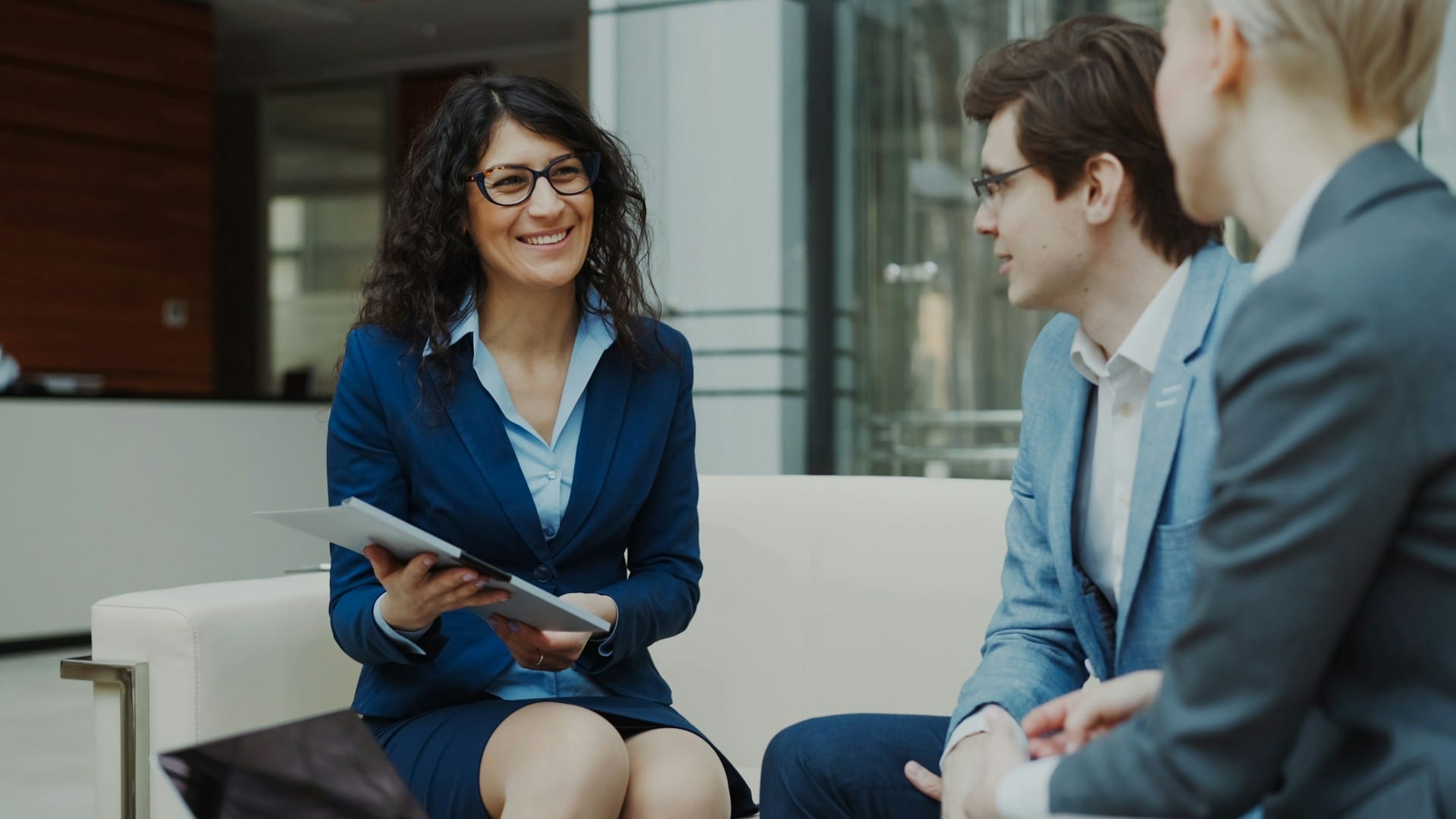 Businesswoman in a navy blazer smiling while discussing documents with two colleagues in a modern office lobby